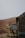 A rustic stone building with a dark, weathered appearance partially visible on the right. Next to it, a sign is mounted, displaying information about Dalehead Bunkhouse, indicating accommodations for 20 people and providing a contact number for booking. The background features a blurred landscape with earthy tones, likely hills or fields, and some out-of-focus trees.