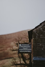 A rustic stone building with a dark, weathered appearance partially visible on the right. Next to it, a sign is mounted, displaying information about Dalehead Bunkhouse, indicating accommodations for 20 people and providing a contact number for booking. The background features a blurred landscape with earthy tones, likely hills or fields, and some out-of-focus trees.