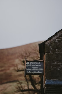 A rustic stone building with a dark, weathered appearance partially visible on the right. Next to it, a sign is mounted, displaying information about Dalehead Bunkhouse, indicating accommodations for 20 people and providing a contact number for booking. The background features a blurred landscape with earthy tones, likely hills or fields, and some out-of-focus trees.