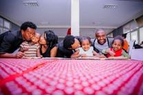 A joyful group of adults and children leaning over a table with a vibrant red tablecloth, smiling warmly at the camera in a well-lit room. The backdrop has multiple windows allowing natural light in, and the people are dressed casually, exuding a sense of togetherness and affection.