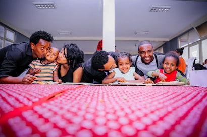 A joyful group of adults and children leaning over a table with a vibrant red tablecloth, smiling warmly at the camera in a well-lit room. The backdrop has multiple windows allowing natural light in, and the people are dressed casually, exuding a sense of togetherness and affection.