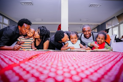 A joyful group of adults and children leaning over a table with a vibrant red tablecloth, smiling warmly at the camera in a well-lit room. The backdrop has multiple windows allowing natural light in, and the people are dressed casually, exuding a sense of togetherness and affection.