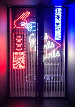 A vibrant display of neon signage behind a mesh screen, featuring colorful elements such as a hand gesture in red, text in both English and another language, and additional abstract shapes and symbols. The signage is illuminated in bright red, blue, and white hues, creating a visually striking and lively atmosphere.