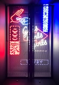 A vibrant display of neon signage behind a mesh screen, featuring colorful elements such as a hand gesture in red, text in both English and another language, and additional abstract shapes and symbols. The signage is illuminated in bright red, blue, and white hues, creating a visually striking and lively atmosphere.