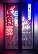A vibrant display of neon signage behind a mesh screen, featuring colorful elements such as a hand gesture in red, text in both English and another language, and additional abstract shapes and symbols. The signage is illuminated in bright red, blue, and white hues, creating a visually striking and lively atmosphere.