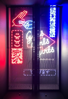 A vibrant display of neon signage behind a mesh screen, featuring colorful elements such as a hand gesture in red, text in both English and another language, and additional abstract shapes and symbols. The signage is illuminated in bright red, blue, and white hues, creating a visually striking and lively atmosphere.