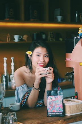 A smiling young woman holding a 'Balik Modal 3,5 Bulan' sign in her bustling coffee shop.