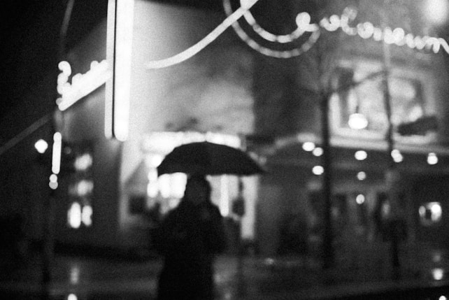 A black and white photograph features a person holding an umbrella on a rainy night in front of a brightly lit, blurred neon sign. The scene conveys a sense of solitude and urban mystery.