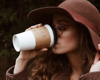 woman with brown hat and tortoiseshell framed eyeglasses drinking coffee in plastic cup