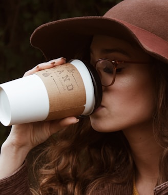 woman with brown hat and tortoiseshell framed eyeglasses drinking coffee in plastic cup