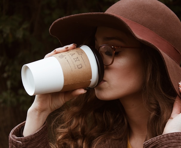 woman with brown hat and tortoiseshell framed eyeglasses drinking coffee in plastic cup