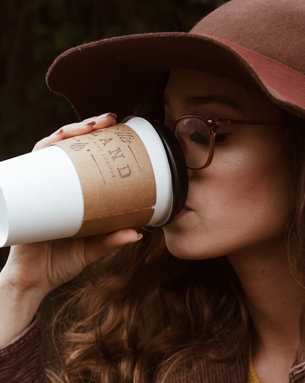 woman with brown hat and tortoiseshell framed eyeglasses drinking coffee in plastic cup