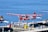 A seaplane with a red and white design featuring a maple leaf is parked at a dock on a calm body of water. The aircraft is labeled with 'HARBOUR AIR' and 'CANADA 150.' Other seaplanes are visible in the background, along with dock equipment such as barrels and crates. The overall setting appears to be a seaplane terminal or harbor.