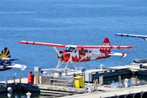 A seaplane with a red and white design featuring a maple leaf is parked at a dock on a calm body of water. The aircraft is labeled with 'HARBOUR AIR' and 'CANADA 150.' Other seaplanes are visible in the background, along with dock equipment such as barrels and crates. The overall setting appears to be a seaplane terminal or harbor.