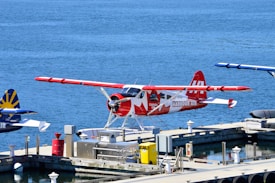 A seaplane with a red and white design featuring a maple leaf is parked at a dock on a calm body of water. The aircraft is labeled with 'HARBOUR AIR' and 'CANADA 150.' Other seaplanes are visible in the background, along with dock equipment such as barrels and crates. The overall setting appears to be a seaplane terminal or harbor.