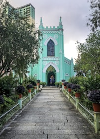 A picturesque pathway leads to a mint-green Gothic-style church with arched windows and a central stained glass feature. The surrounding area is lush with trees and neatly arranged potted plants, creating a serene and inviting atmosphere. In the background, a modern apartment building subtly rises behind the church.