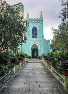 A picturesque pathway leads to a mint-green Gothic-style church with arched windows and a central stained glass feature. The surrounding area is lush with trees and neatly arranged potted plants, creating a serene and inviting atmosphere. In the background, a modern apartment building subtly rises behind the church.