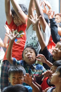 A group of children are enthusiastically raising their hands and making expressive faces, possibly during a game or an interactive activity. The scene is lively and crowded, with the children wearing colorful clothes. There is a sense of movement and excitement among the group.