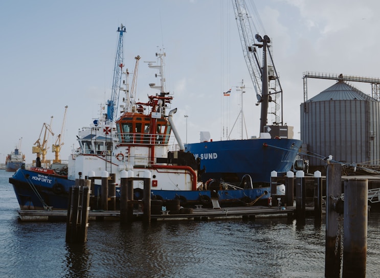 Several industrial ships are docked at a port, surrounded by various cranes and large storage silos. The water is calm, and there is a clear view of the vessels' structures and equipment.