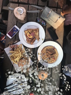 A rustic wooden table is set with a variety of brunch items including a plate of waffles topped with fried chicken, a plate of toast with sausages and orange wedges, and a burger with fries. A whole coconut with a straw is also present. There are two smartphones, a pair of sunglasses, and a purse on the table. Decorative white flowers are placed in the foreground.
