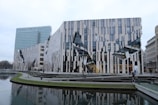 A modern architectural building with a distinctive facade of glass and striped panels, incorporating sharp angles and geometric shapes. The foreground features a reflective water canal and pathways with patches of grass. Other urban buildings are visible in the background, showcasing a blend of classic and contemporary architecture.