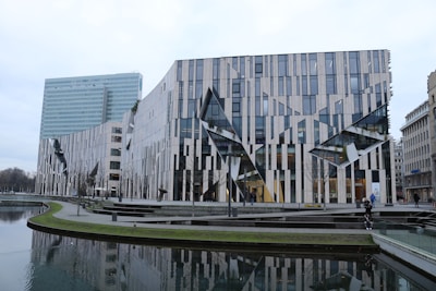 A modern architectural building with a distinctive facade of glass and striped panels, incorporating sharp angles and geometric shapes. The foreground features a reflective water canal and pathways with patches of grass. Other urban buildings are visible in the background, showcasing a blend of classic and contemporary architecture.