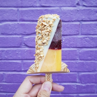 A hand is holding a colorful popsicle against a purple brick wall. The popsicle is divided into three sections: one part is coated with granola, another is a dark berry color, and the bottom is a light orange hue.