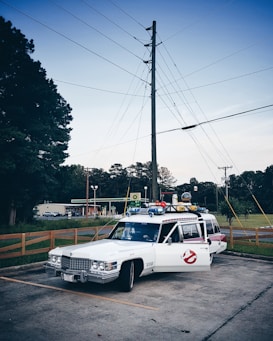A vintage white Cadillac hearse is parked in an outdoor setting, adorned with various gadgets and logos reminiscent of the Ghostbusters vehicle. It is parked near a gasoline station, with grassy areas and large trees in the background. Overhead, power lines and a pale blue sky are visible.