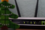 A close-up view of a Wi-Fi router with various indicator lights lit up, showing connectivity status. To the left, the blurred image of a decorative object, resembling a green Christmas tree, is partially visible.
