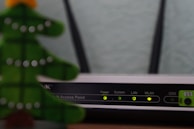 A close-up view of a Wi-Fi router with various indicator lights lit up, showing connectivity status. To the left, the blurred image of a decorative object, resembling a green Christmas tree, is partially visible.