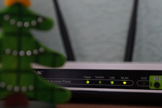 A close-up view of a Wi-Fi router with various indicator lights lit up, showing connectivity status. To the left, the blurred image of a decorative object, resembling a green Christmas tree, is partially visible.
