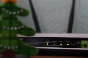 A close-up view of a Wi-Fi router with various indicator lights lit up, showing connectivity status. To the left, the blurred image of a decorative object, resembling a green Christmas tree, is partially visible.