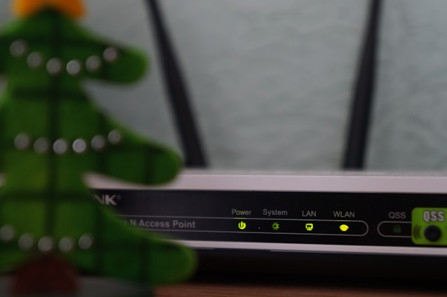 A close-up view of a Wi-Fi router with various indicator lights lit up, showing connectivity status. To the left, the blurred image of a decorative object, resembling a green Christmas tree, is partially visible.