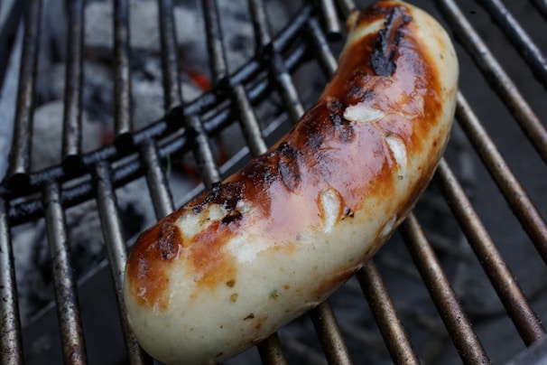 A grilled sausage with a crispy, browned exterior rests on metal grates, with char marks visible on its surface. The background shows part of a charcoal grill.