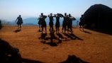 A group of people dressed in traditional attire perform a dance on an earthy, open area with mountains in the background. The lighting creates distinct shadows, and a round hut structure is visible on the right.