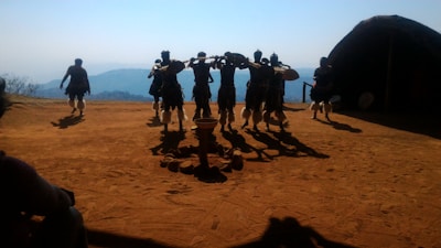 A group of people dressed in traditional attire perform a dance on an earthy, open area with mountains in the background. The lighting creates distinct shadows, and a round hut structure is visible on the right.