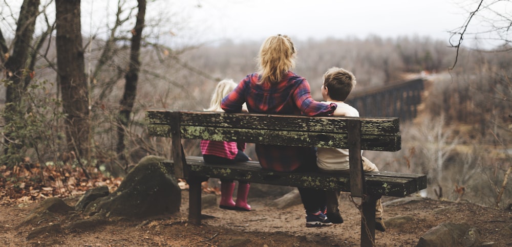 donna tra due bambini seduta su panca di legno marrone durante il giorno