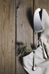 A wooden table surface with a textured appearance, featuring a silver cake server and fork placed on a white fabric napkin. Accompanying the utensils is a sprig of rosemary with a small purple berry, adding a touch of nature to the arrangement.