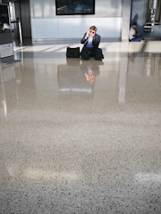 A man in formal attire sits on a shiny, speckled floor, talking on the phone. He has a bag beside him and wears sunglasses. In the background, another person sits against the wall, possibly using a phone. The setting appears to be a modern public space or airport terminal with large windows allowing natural light to create reflections on the floor.
