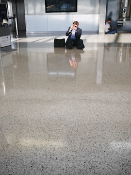 A man in formal attire sits on a shiny, speckled floor, talking on the phone. He has a bag beside him and wears sunglasses. In the background, another person sits against the wall, possibly using a phone. The setting appears to be a modern public space or airport terminal with large windows allowing natural light to create reflections on the floor.