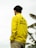 A person is wearing a bright yellow jacket with 'Erigo Beach Club' printed on the back. In the background, there are leaves, possibly from banana trees, against an overcast sky.