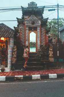 A person in casual attire stands at the entrance of an ornate temple gate, featuring intricate carvings and a large mirror or window reflecting lush green scenery. The setting appears to be on a street, with lampposts and power lines framing the view. The atmosphere is serene and cultural, with decorative statues flanking the entrance.