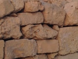 Rough, irregularly shaped stones stacked in a formation resembling a wall. The stones have a natural, uneven texture and are primarily a light brown color with some darker shading. Small gaps are visible between some of the stones.