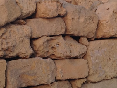 Rough, irregularly shaped stones stacked in a formation resembling a wall. The stones have a natural, uneven texture and are primarily a light brown color with some darker shading. Small gaps are visible between some of the stones.