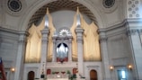 An ornate and grand interior of a building featuring an impressive pipe organ with tall golden pipes set against a backdrop of a circular and intricately decorated ceiling. The architecture includes detailed moldings and columns, creating a sense of majesty and elegance.