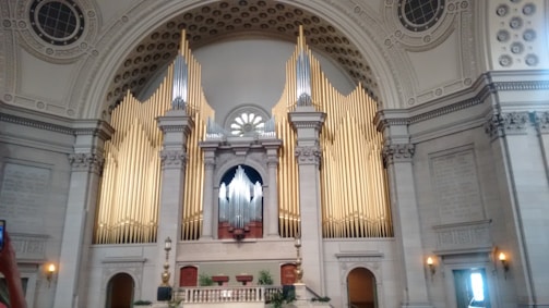 An ornate and grand interior of a building featuring an impressive pipe organ with tall golden pipes set against a backdrop of a circular and intricately decorated ceiling. The architecture includes detailed moldings and columns, creating a sense of majesty and elegance.