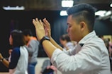 A group of people appear engaged in an activity that involves raising hands. The central person in focus is a man with short hair, wearing a white shirt and a black wristwatch, seemingly in concentration or worship.
