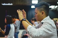A group of people appear engaged in an activity that involves raising hands. The central person in focus is a man with short hair, wearing a white shirt and a black wristwatch, seemingly in concentration or worship.