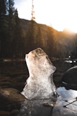 A block of ice stands upright in a shallow stream surrounded by smooth stones. The background features a forest with tall pine trees and a mountain silhouette under a brightly shining sun, casting warm light and long shadows.