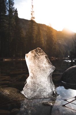 A block of ice stands upright in a shallow stream surrounded by smooth stones. The background features a forest with tall pine trees and a mountain silhouette under a brightly shining sun, casting warm light and long shadows.